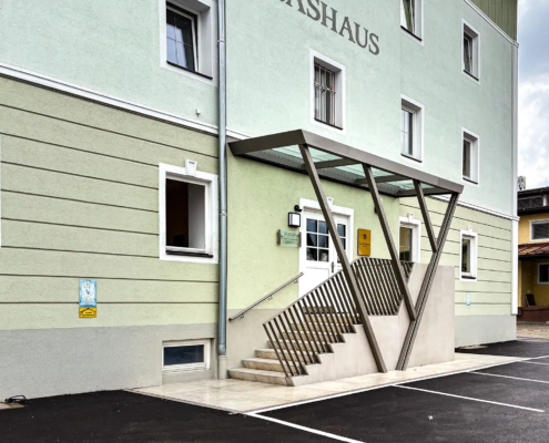 Stahlvordachkonstruktion über einer Eingangsstiege in ein Haus mit einer hellgrünen Fassade. Das Vordach ist aus Glas. Das Geländer ist grau lackiert und der Handlauf ist aus Edelstahl.