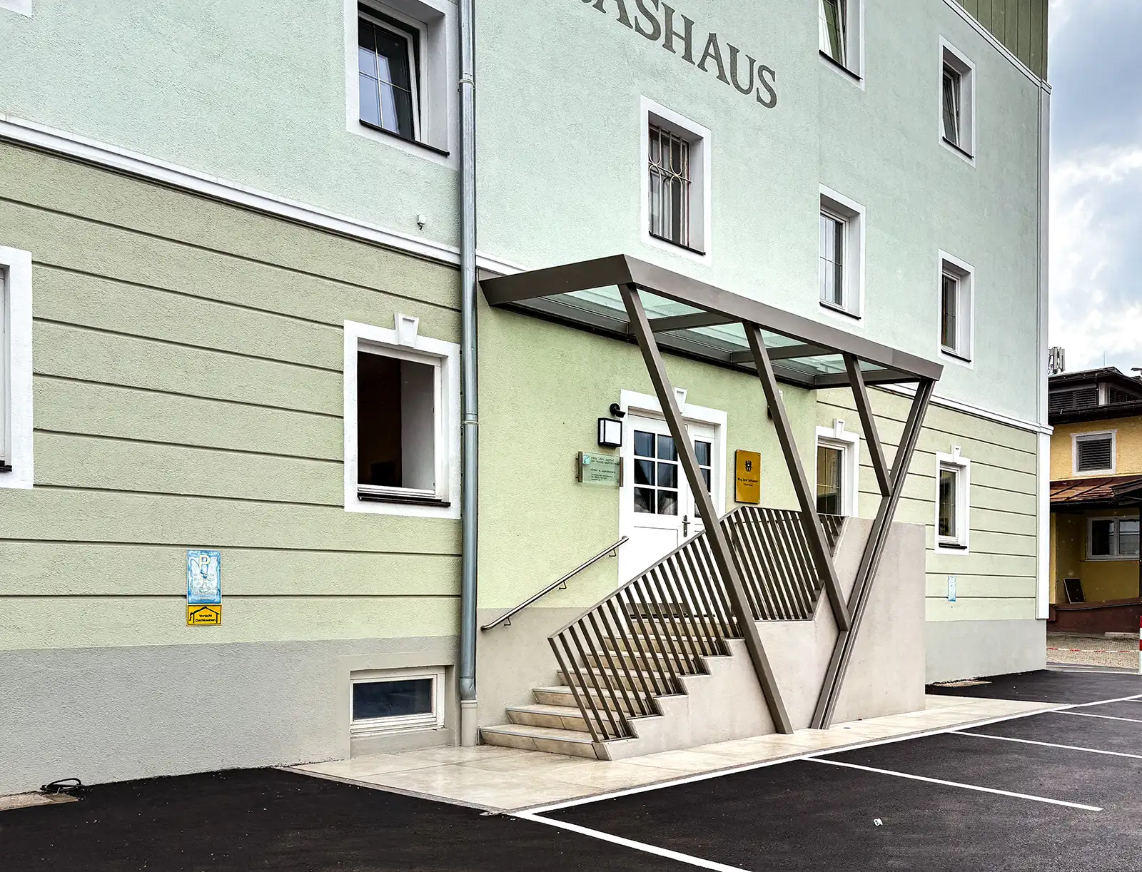 Stahlvordachkonstruktion über einer Eingangsstiege in ein Haus mit einer hellgrünen Fassade. Das Vordach ist aus Glas. Das Geländer ist grau lackiert und der Handlauf ist aus Edelstahl.