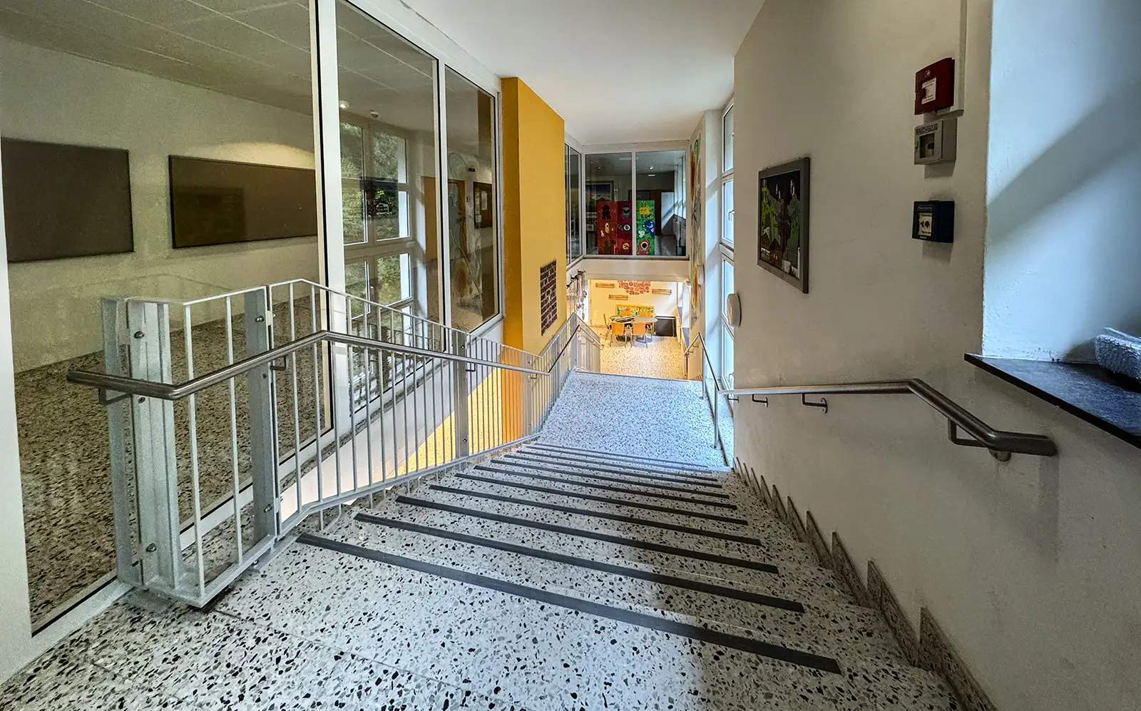 Metallgeländer an einer Stiege in einem Stiegenhaus einer Schule. Detailaufnahme der Stiege mit dem Blick vom ersten Stock nach unten. Das Geländer ist grau lackiert, der Handlauf ist aus Edelstahl. Inder Mitte der Stiege ist eine orange Wand mit einem Kunstwerk.