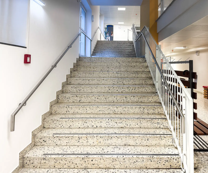 Metallgeländer an einer Stiege in einem Stiegenhaus einer Schule. Detailaufnahme der Stiege mit dem Blick in den ersten Stock. Das Geländer ist grau lackiert, der Handlauf ist aus Edelstahl.