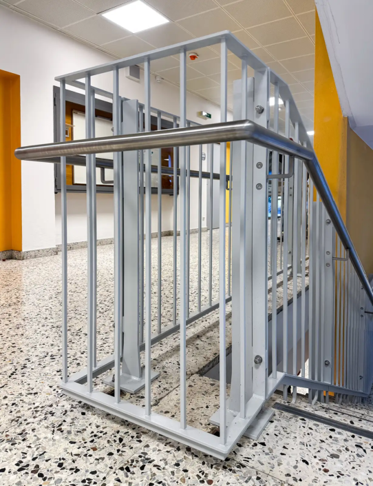 Metallgeländer an einer Stiege in einem hell beleuchteten Stiegenhaus in einer Schule. Detailaufnahme im ersten Stock. Das Geländer ist hell grau lackiert, der Handlauf ist aus Edelstahl