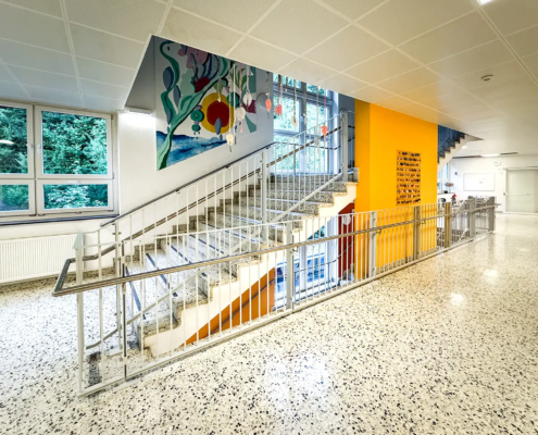 Metallgeländer an einer Stiege in einem hell beleuchteten Stiegenhaus in einer Schule. Einer orange Wand mit einem Kunstwerk ist in der Mitte der Stiege und im Hintergrund schmückt ein Gemälde die Wand
