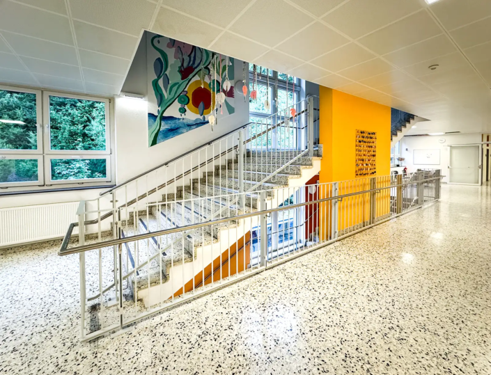 Metallgeländer an einer Stiege in einem hell beleuchteten Stiegenhaus in einer Schule. Einer orange Wand mit einem Kunstwerk ist in der Mitte der Stiege und im Hintergrund schmückt ein Gemälde die Wand