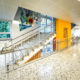 Metallgeländer an einer Stiege in einem hell beleuchteten Stiegenhaus in einer Schule. Einer orange Wand mit einem Kunstwerk ist in der Mitte der Stiege und im Hintergrund schmückt ein Gemälde die Wand