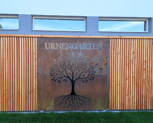 Urnengarten Jochberg, mit einem Laser ausgeschnittener Bauch in einem rostigen Corten-Stahl-Blech. Links und rechts ist eine Wand aus Holzlatten