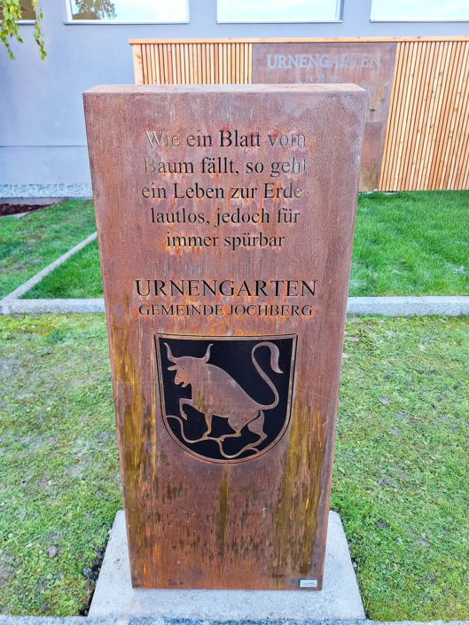 Säule aus einem rostigen Corten-Stahl Blech mit einem Wappen der Gemeinde Jochberg und einem Text mit der Hauptüberschrift "Urnengarten"